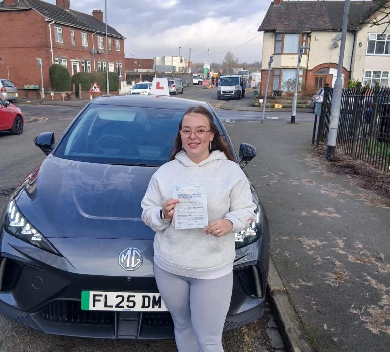 A person stands next to a grey car, holding a certificate on a residential street.