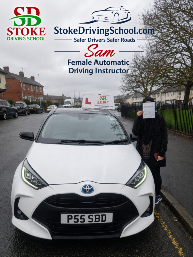 A person holding a sign stands beside a white car with learner plates on a street.
