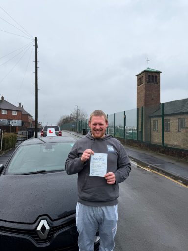 A man holds a certificate, standing next to a driving school car on a cloudy street.