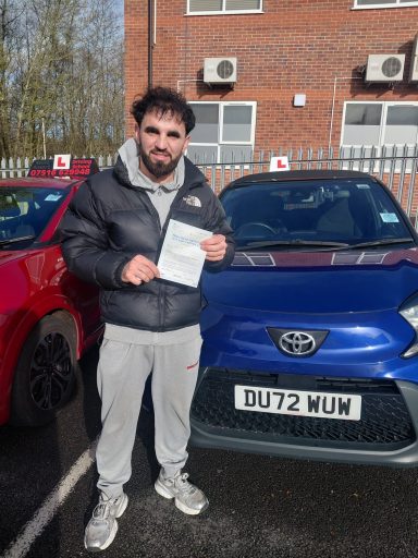 Man in a black jacket holds a document next to a blue Toyota car.