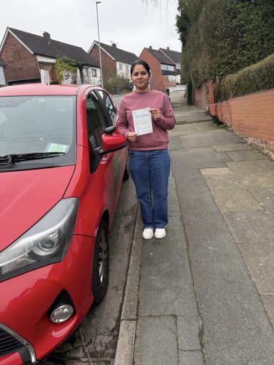 A person stands next to a red car holding a document on a residential street.