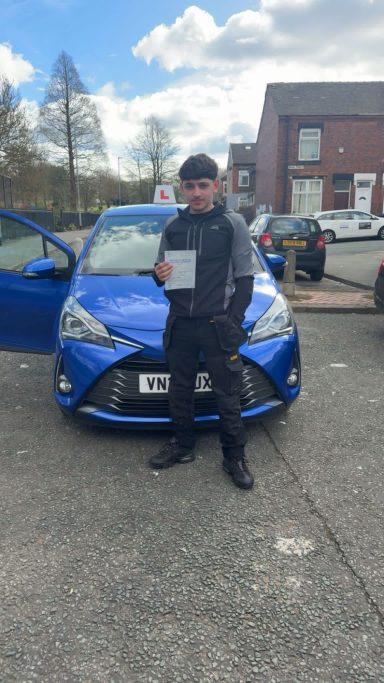 A person stands next to a blue car, holding a sign that says "L" for learner driver.