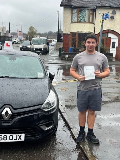 A young man stands beside a parked black Renault car, holding a driving certificate.