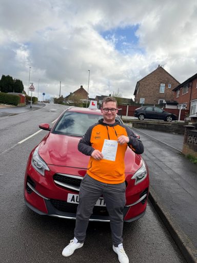 Person in an orange top holds a driving certificate in front of a red car on a street.