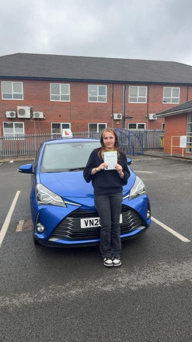 A young woman holds a driving test certificate in front of a blue car.