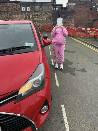 A person in a pink tracksuit stands by a red car, holding something in front of their face.