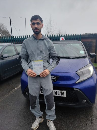 A young man in a tracksuit holds a certificate in front of a blue car.