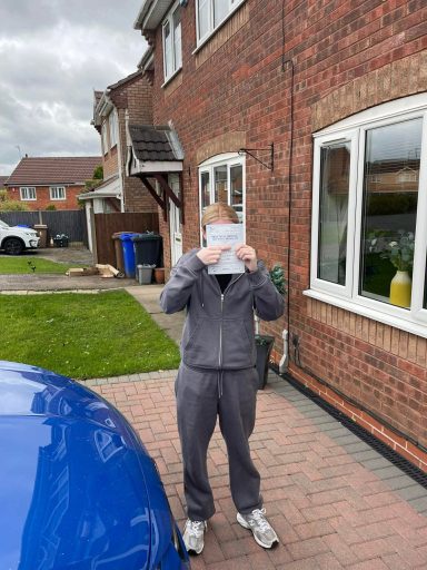 A person in grey clothing holds a piece of paper in front of their face outside a house.