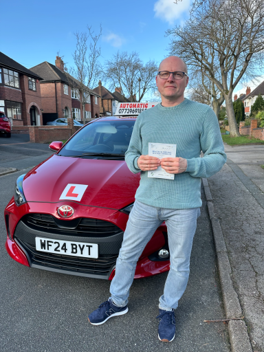 A man stands beside a red learner driver car, holding a driving theory test certificate.