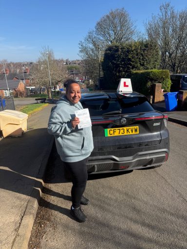 A learner driver holds a certificate in front of a car with "L" plates.