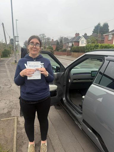 A woman holding papers stands next to an open car door on a cloudy day.
