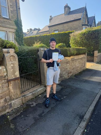 A person stands on a street holding a document, with hedges and houses in the background.