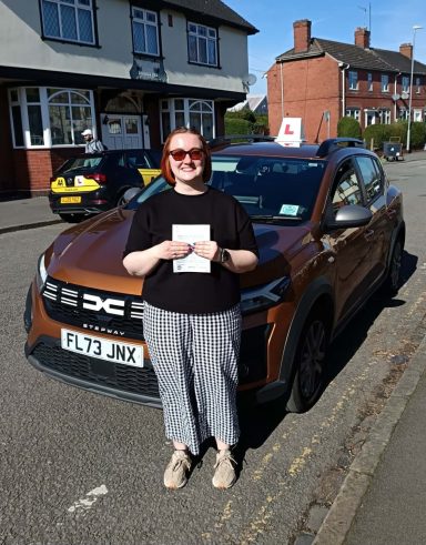 A woman stands next to a brown car with a learner's sign in a residential area.