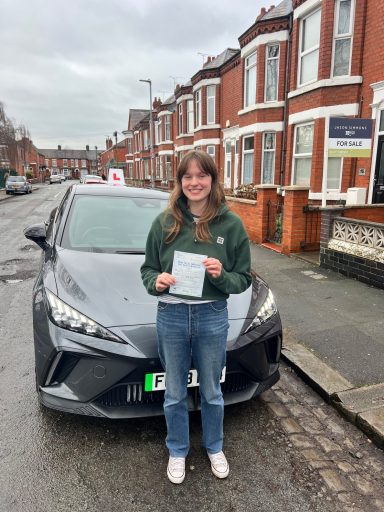 A young woman stands by a grey car, holding a driving test certificate on a residential street.
