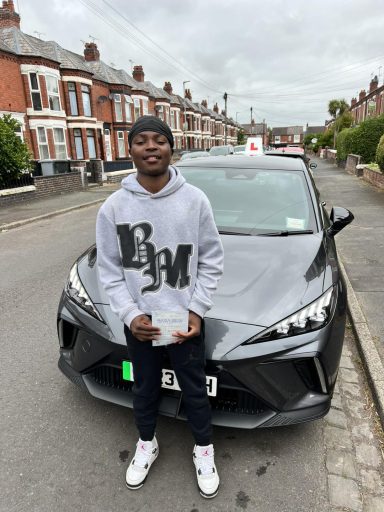 A young man in a grey hoodie stands next to a silver car with a learner driver sign.