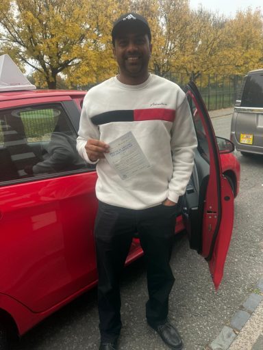 A man stands by a red car, holding a document, with autumn trees in the background.