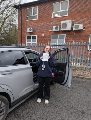 A person holding a certificate stands next to a grey car, with a building and air conditioning units behind.