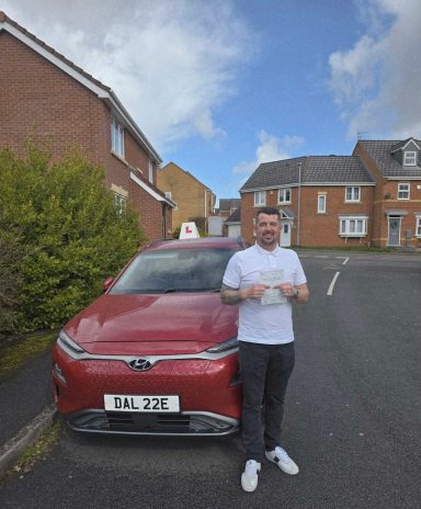 A man stands beside a red car displaying a learner driver sign, holding a certificate.