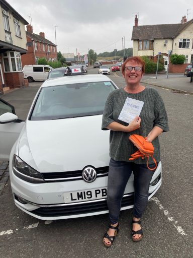 Smiling person holding paperwork stands beside a white Volkswagen car in a residential area.
