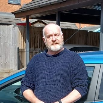 Bearded man in a blue sweater standing next to a car, with a wooden fence in the background.