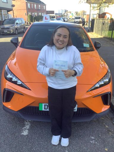A smiling person holds a certificate in front of an orange car.