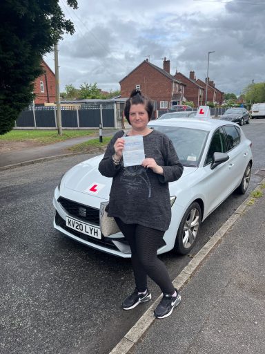 A woman stands next to a white car, holding a certificate on a residential street.