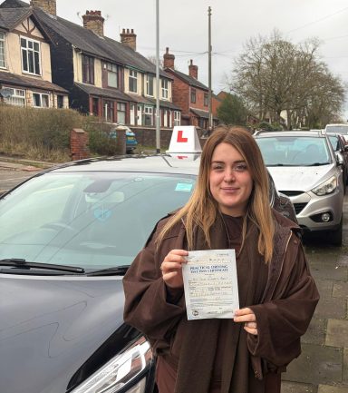 A person holding a driving test certificate stands near a car with a learner plate.