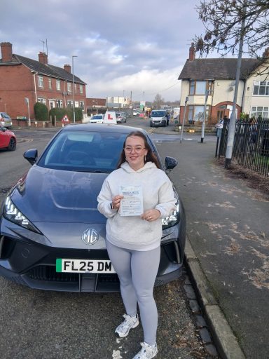 A person holding a document stands beside a grey car on a street.