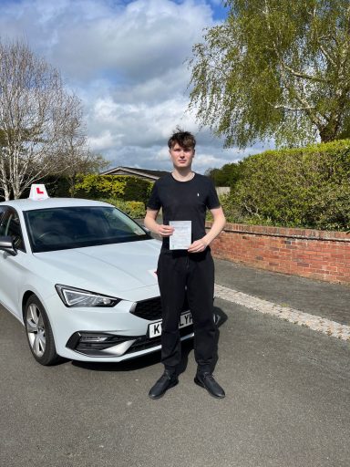 A young man stands beside a car, holding a driving test certificate.