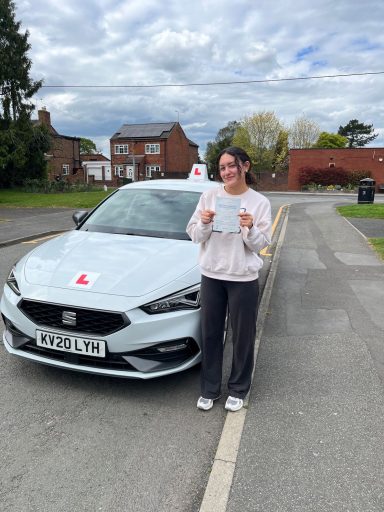 A woman stands with a driving certificate next to a learner driver car on a suburban street.
