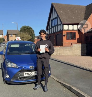 A young man holds a certificate next to a blue car parked on a residential street.