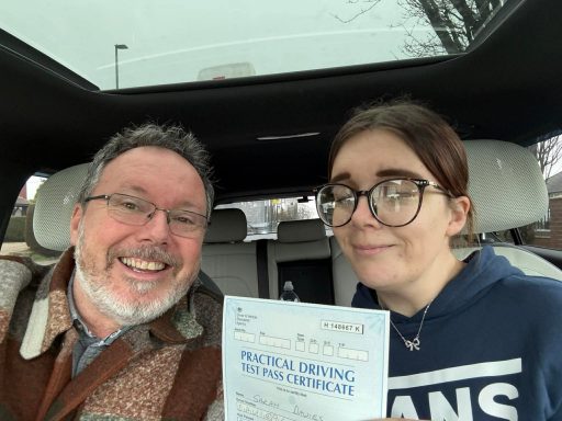 Two people inside a car, one smiles widely while holding a document, the other looks unsure.
