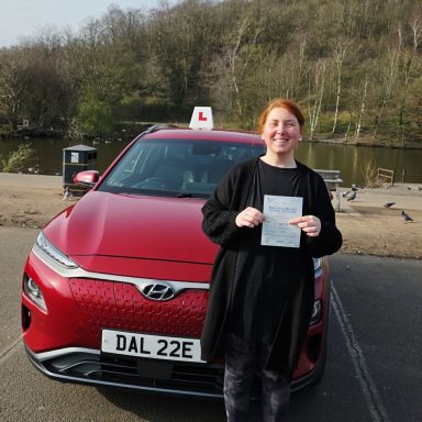A woman holds a certificate in front of a red driving instructor car by a lake.
