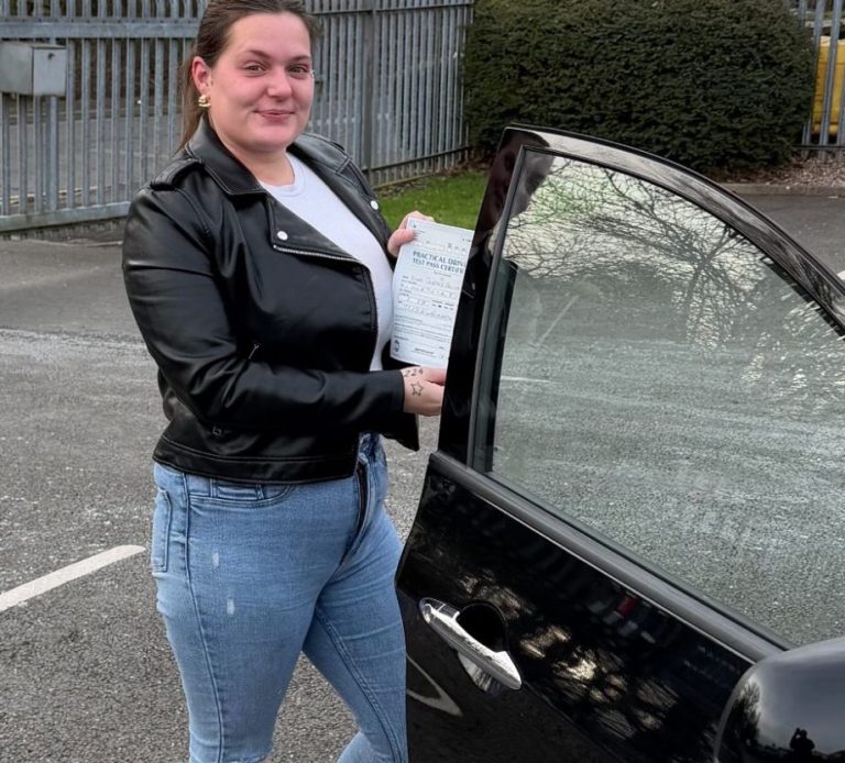 A woman in a black leather jacket stands next to an open car door, holding documents.