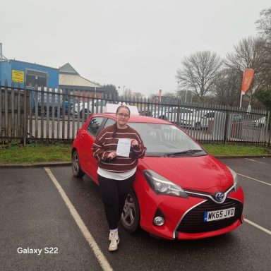 A person holding a document stands beside a red car in a parking area.