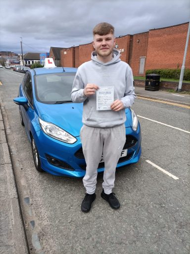 A person in casual attire stands beside a blue car, holding a certificate.