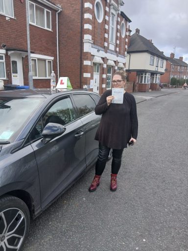 A person wearing black holding a paper stands beside a grey car on a street.