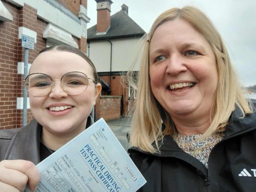 Two women smiling outdoors, holding a ticket or document in front of a building.