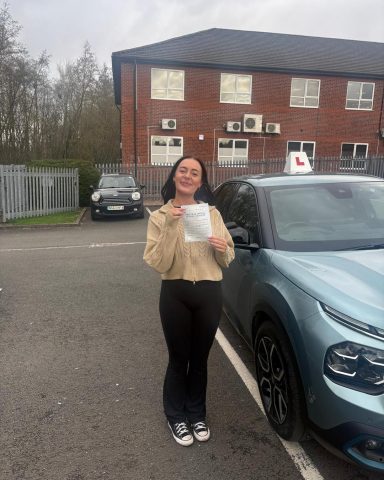 A woman stands beside a blue car, holding a certificate and smiling.