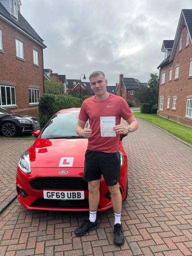 A young man in a red t-shirt poses with a driving certificate next to a red car.