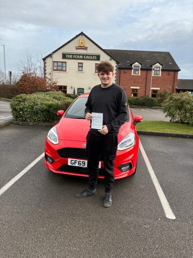 Young man holding a certificate stands in front of a red car parked outside a pub.