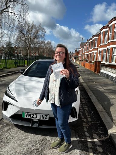 A woman stands beside a white car, holding a document on a sunny street.