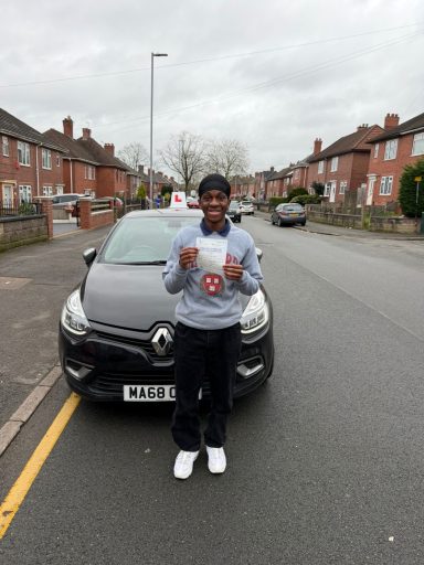 A young person holds a driving test pass certificate in front of a car on a residential street.
