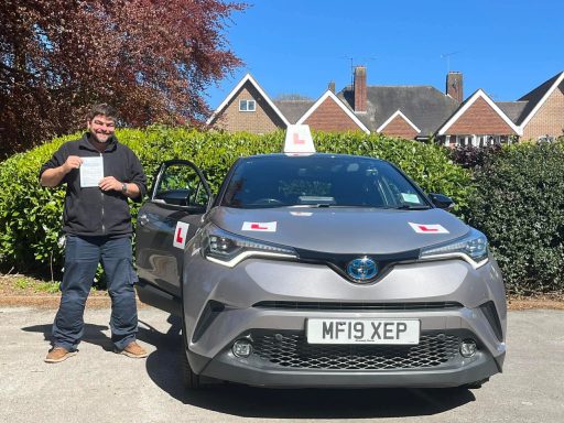 A man stands next to a grey car with a driving school sign, holding a certificate.