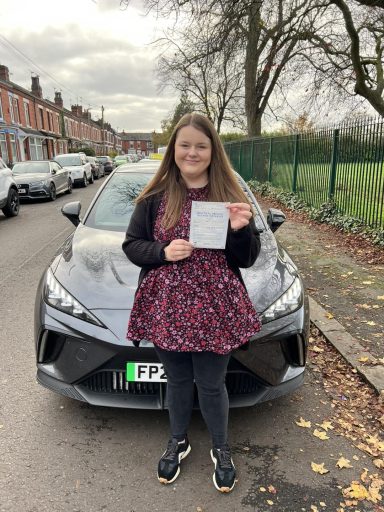 A young woman in a floral dress holding a paper outside a parked car on a street.