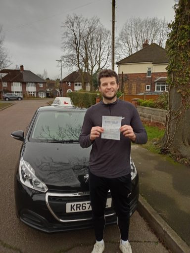 A young man holds a driving certificate beside a taxi in a residential area.