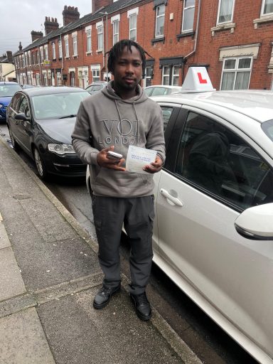 A person in casual attire stands next to a learner driver car holding a document.