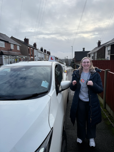 A young person stands beside a car, smiling, in a residential area with cloudy skies.