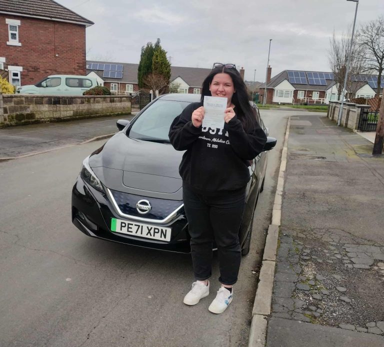 A person in a black hoodie holds a document beside a black car on a residential street.