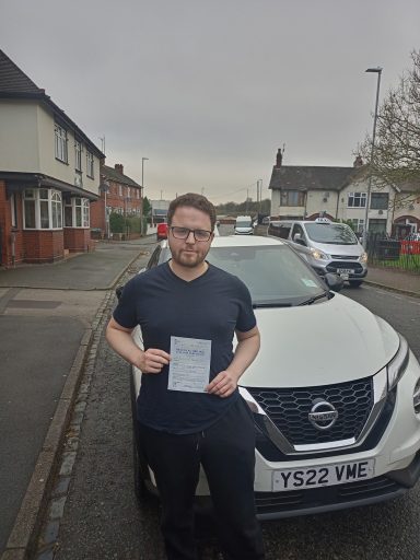 A man holding a document stands in front of a white Nissan car on a suburban street.